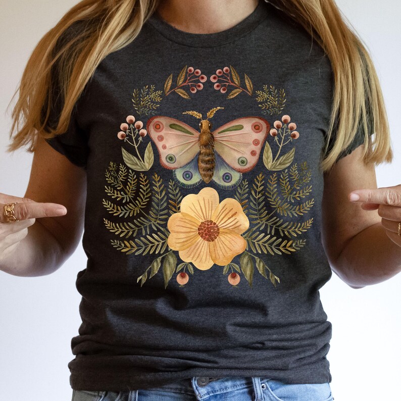 Pu&ograve; includere: T-shirt grigio scuro con un disegno ad acquerello di una farfalla, un fiore e un fogliame. La farfalla ha ali rosa, blu e marroni. Il fiore &egrave; giallo con un centro rosso. Il disegno &egrave; circondato da foglie verdi e bacche.