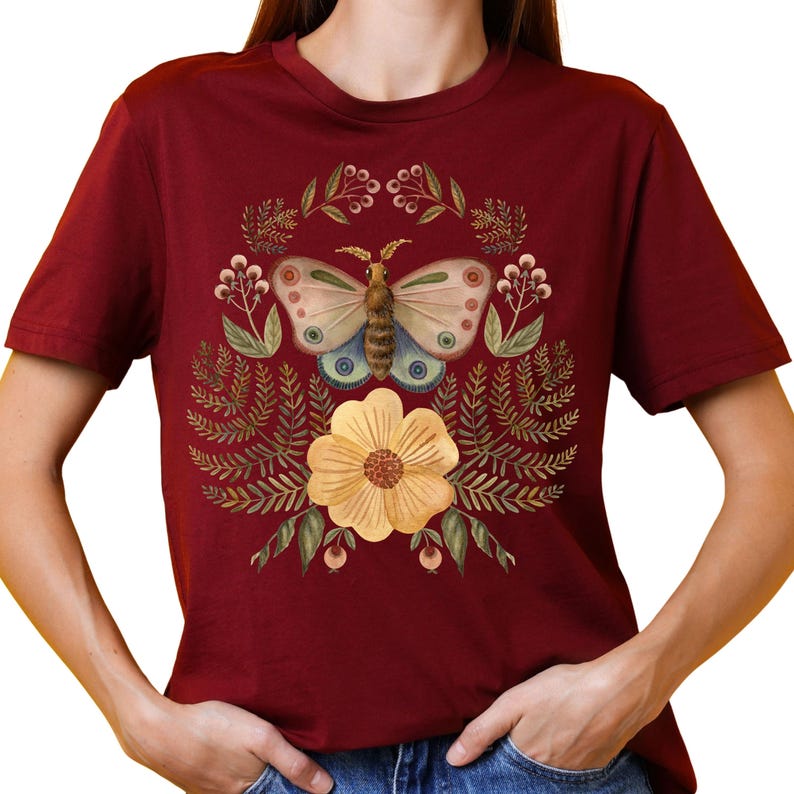 Pu&ograve; includere: T-shirt bordeaux con un disegno ad acquerello di una farfalla, un fiore giallo e foglie. La farfalla ha ali azzurre e beige con dettagli marroni e blu. Il disegno &egrave; centrato sulla maglietta.