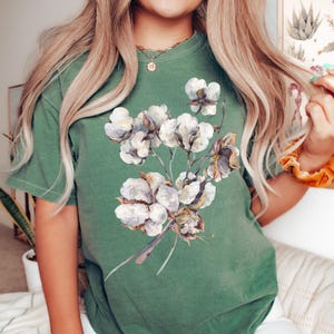 May include: A forest green short-sleeve t-shirt featuring a central graphic print of a cluster of white cotton bolls with brown stems and husks. The cotton illustration displays soft, watercolour-style shading and detailed textures on the petals.