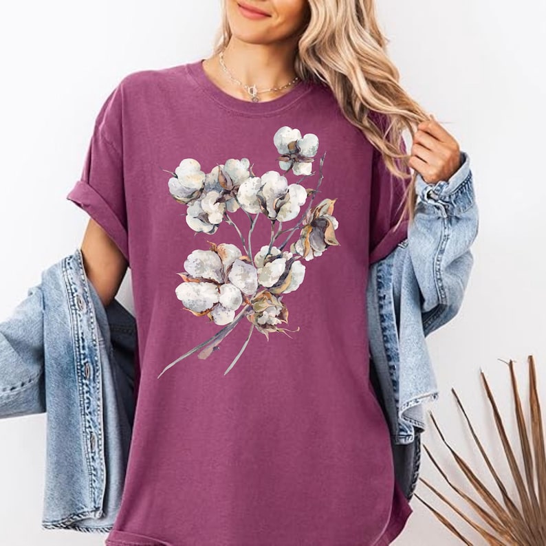 May include: A dark mauve short-sleeved t-shirt featuring a central graphic print of a cluster of white cotton bolls on brown stems. The shirt is worn with a light blue denim jacket draped over the shoulders, showcasing a casual, layered appearance.