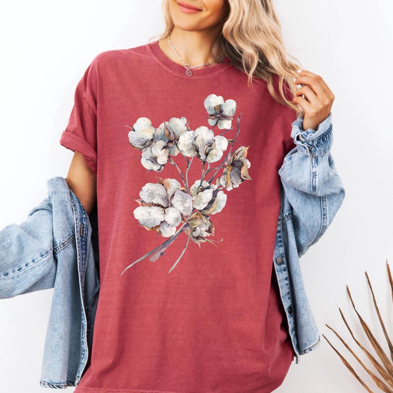 May include: A short-sleeved, crew-neck t-shirt in a muted, washed-out red colour. The front of the shirt features a detailed, watercolour-style illustration of a cluster of white cotton bolls with brown stems and husks centred on the chest.