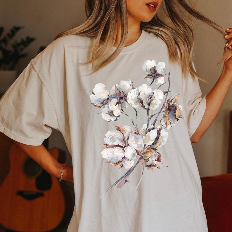 May include: A beige short-sleeved t-shirt featuring a central graphic print of a cotton plant branch. The illustration displays multiple fluffy white cotton bolls with brown husks and thin dark stems, rendered in a soft watercolour style.