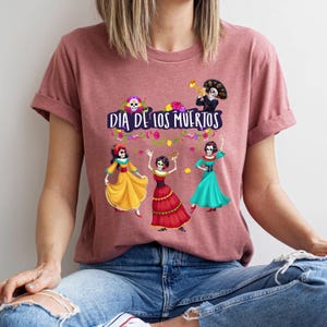 Op de afbeelding: Een mauve t-shirt met korte mouwen en een grafische print van drie dansende skeletten in kleurrijke jurken en &eacute;&eacute;n skelet dat trompet speelt. De tekst "DIA DE LOS MUERTOS" staat in het wit boven de figuren, omringd door bloemaccenten.