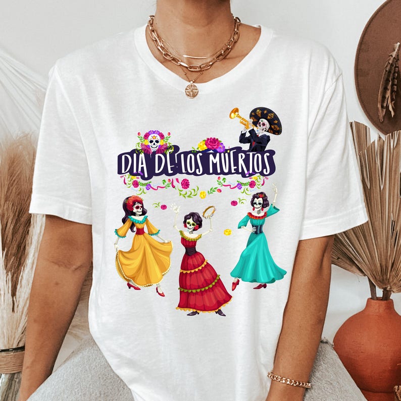 Op de afbeelding: Een wit t-shirt met een kleurrijke afbeelding van vier skeletfiguren in traditionele kleding, met de tekst "DIA DE LOS MUERTOS" in het paars gedrukt. Het ontwerp bevat bloemaccenten en een skelet dat een trompet bespeelt.