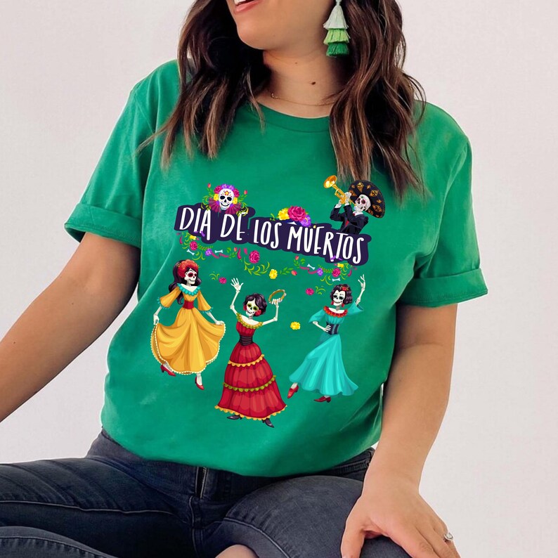 Op de afbeelding: Een felgroen T-shirt met korte mouwen en een grafische print van drie dansende skeletten in kleurrijke jurken en &eacute;&eacute;n skelet dat trompet speelt. De tekst "DIA DE LOS MUERTOS" staat in een donkerpaars lettertype boven de figuren.
