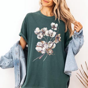 May include: A dark forest green short-sleeve t-shirt featuring a central graphic print of a cluster of white cotton bolls with brown stems and leaves. The shirt is worn with a light blue denim jacket draped over the shoulders, showing rolled sleeves.