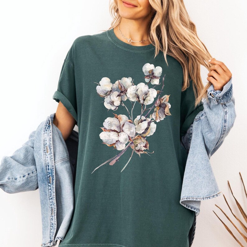 May include: A dark forest green short-sleeve t-shirt featuring a central graphic print of a cluster of white cotton bolls with brown stems and leaves. The shirt is worn with a light blue denim jacket draped over the shoulders, showing rolled sleeves.
