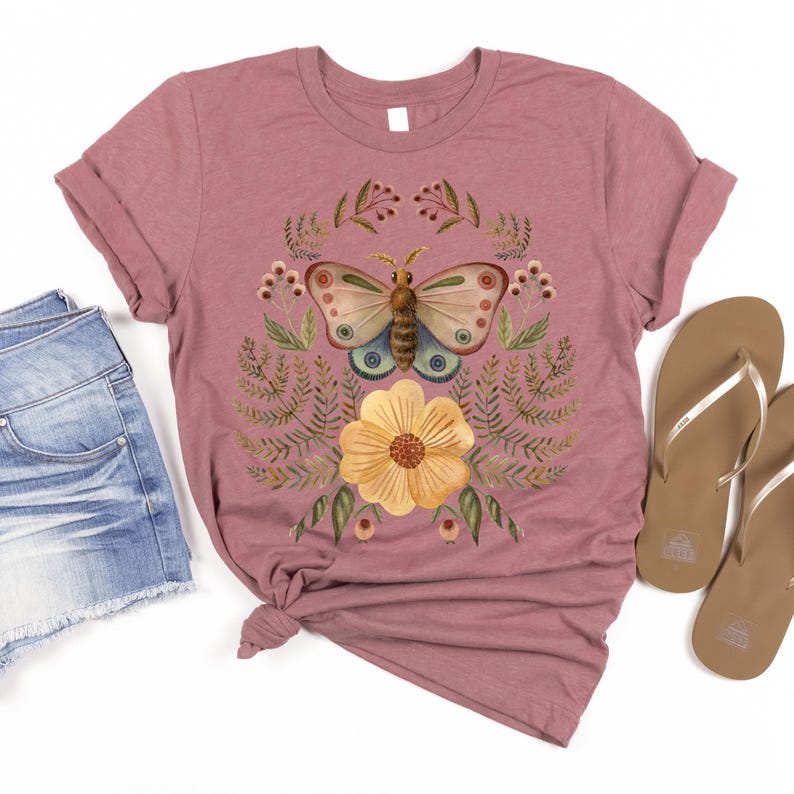 Pu&ograve; includere: T-shirt rosa antico con un disegno di farfalla ad acquerello, un fiore giallo e accenti botanici. La farfalla ha ali blu, marroni e crema. La maglietta &egrave; abbinata a pantaloncini di jeans e infradito marroni.