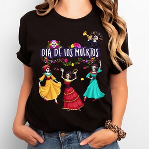 Op de afbeelding: Een zwart t-shirt met een grafische print met de tekst "DIA DE LOS MUERTOS" in het wit. Het ontwerp bevat vier skeletfiguren gekleed in kleurrijke traditionele kleding, omringd door bloemaccenten en een trompetspeler.