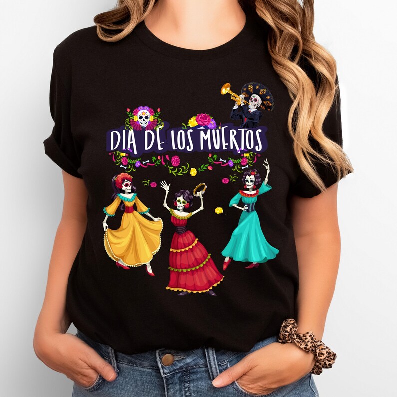 Op de afbeelding: Een zwart t-shirt met een grafische print met de tekst "DIA DE LOS MUERTOS" in het wit. Het ontwerp bevat vier skeletfiguren gekleed in kleurrijke traditionele kleding, omringd door bloemaccenten en een trompetspeler.