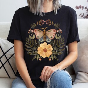 Pu&ograve; includere: T-shirt nero con un design a farfalla e floreale. La farfalla ha dettagli beige, rosa e blu, circondata da un fiore giallo e fogliame verde. La maglietta &egrave; a girocollo e maniche corte.