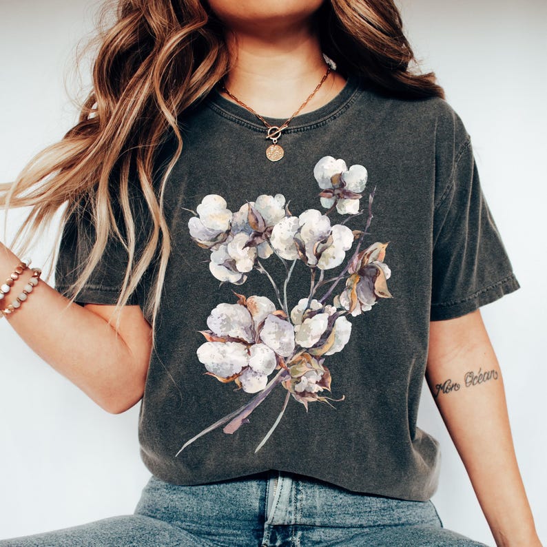 May include: A charcoal grey short-sleeve t-shirt featuring a central graphic illustration of a cluster of white cotton bolls on thin brown stems. The shirt has a crew neckline and a vintage-style washed finish, showcasing a botanical design.