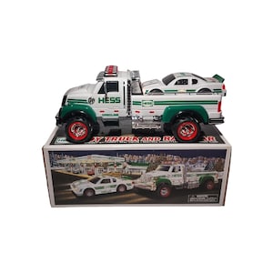 May include: A white and green toy truck with a race car on the back, sitting on a box. The truck has red wheels and a red light bar. The box has the words "HESS" and "TOY TRUCK AND RACE CAR" printed on it.