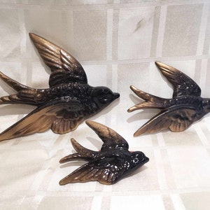 May include: Three ceramic wall decor swallows in black and gold. The largest swallow is in the centre and the two smaller swallows are on either side.