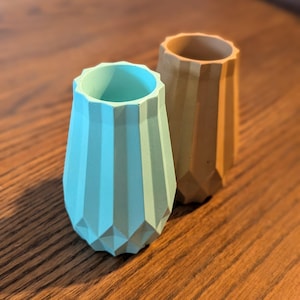 May include: Two geometric concrete vases in a faceted design. One is teal, the other tan. They have a cylindrical shape, suitable for small plants or decorative items. Each vase is approximately 10 cm tall.