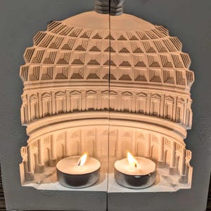 May include: White ceramic candle holder shaped like a dome with two lit tea candles inside. The dome has a detailed design of a building with columns and arches.