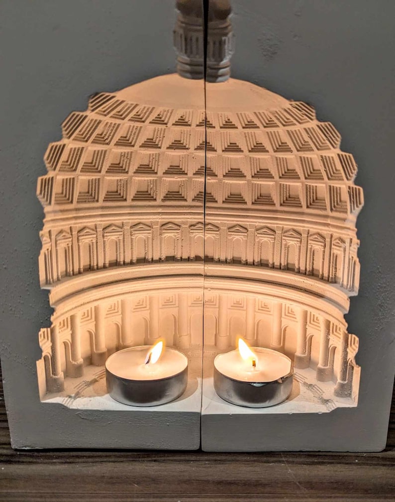 May include: White ceramic candle holder shaped like a dome with two lit tea candles inside. The dome has a detailed design of a building with columns and arches.