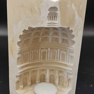 May include: A white plaster candle holder shaped like a building with a dome. The candle holder has a small, round opening for a tea light candle.