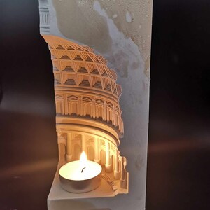 May include: A white plaster candle holder shaped like a Roman building with a lit tea light candle inside.
