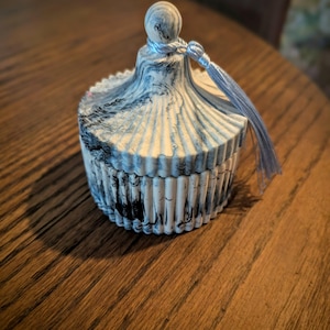 May include: A decorative trinket box with a lid. The box is white with black and blue marbling. The lid has a small ball on top and a blue tassel. The box is on a wooden table.