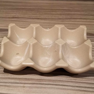 May include: A beige ceramic egg holder with six individual compartments. The holder has a wavy design and is made of a speckled material. The egg holder is designed to hold six eggs.