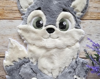 May include: A plush, gray wolf-shaped pillow with white accents and large green eyes. The wolf has a smiling expression and paw-shaped details on its feet. The pillow is set against a wooden background.