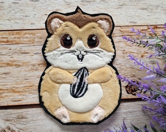 May include: A brown and white plush hamster holding a black and white sunflower seed. The hamster is embroidered with black eyes and a black nose. The hamster is sitting on a wooden surface.