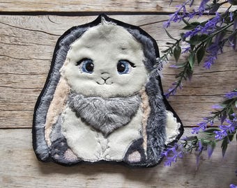 May include: A gray and white embroidered bunny patch with blue eyes. The patch is made of soft, plush fabric and has a fuzzy texture.