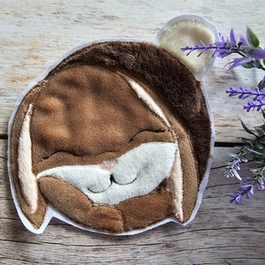 Könnte beinhalten: Ein braunes Plüschkaninchen mit weißen Ohren und einer weißen Nase, das mit geschlossenen Augen lächelt. Das Kaninchen ist auf einen weißen Stoffhintergrund genäht.