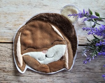 Könnte beinhalten: Ein braunes Plüschkaninchen mit weißen Ohren und einer weißen Nase, das mit geschlossenen Augen lächelt. Das Kaninchen ist auf einen weißen Stoffhintergrund genäht.