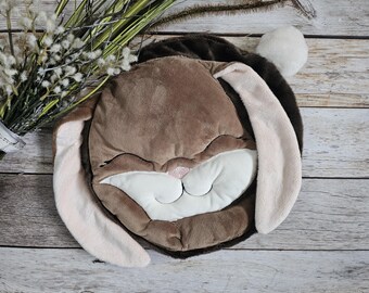 Könnte beinhalten: Ein braunes Plüsch-Hasenkissen mit einem lächelnden Gesicht und rosa Ohren. Das Kissen liegt auf einer Holzoberfläche.