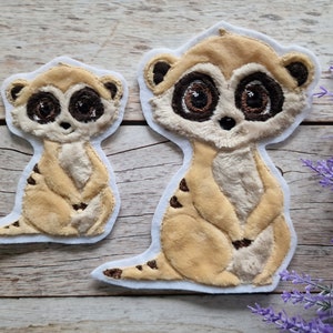 Könnte beinhalten: Zwei gestickte Filzpatches von Erdmännchen. Das größere Erdmännchen sitzt mit gekreuzten Pfoten und hat eine hellbraune Fellfarbe. Das kleinere Erdmännchen steht mit zusammengefügten Pfoten und hat eine dunkelbraune Fellfarbe. Beide Erdmännchen haben schwarze Augen und Nasen.