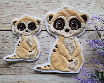 May include: Two embroidered felt patches of meerkats. The larger meerkat is sitting with its paws crossed and has a light brown fur color. The smaller meerkat is standing with its paws together and has a darker brown fur color. Both meerkats have black eyes and noses.