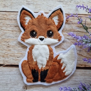 Op de afbeelding: Een bruine en witte pluche vos-applique met zwarte ogen en een witte buik. De vos zit met zijn staart om zijn lichaam gekruld.