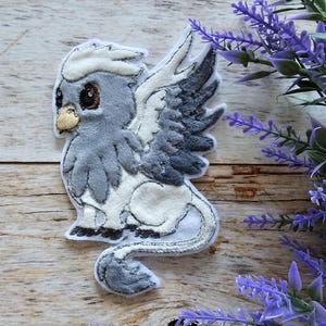 May include: A gray and white embroidered patch of a mythical creature with wings, a bird-like head, and a lion-like body. The creature has large, round eyes and a fluffy tail.