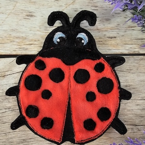 Könnte beinhalten: Ein roter Marienkäfer mit schwarzen Punkten und einem schwarzen Kopf mit weißen Augen. Der Marienkäfer ist aus Plüsch gefertigt und hat ein gesticktes Design.