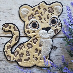 Könnte beinhalten: Ein Stoffaufnäher in Form eines Cartoon-Leopardenbabys mit cremefarbenem Fell, braunen Flecken und großen dunklen Augen. Das Design hat einen schwarzen gestickten Rand, eine weiße Schnauze und einen langen geschwungenen Schwanz in weicher Optik.