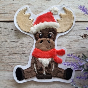 Könnte beinhalten: Ein brauner Plüsch-Elch, der einen roten Weihnachtsmann-Hut und einen roten Schal trägt. Der Elch sitzt mit gekreuzten Beinen und hat einen weißen Fleck auf der Brust.