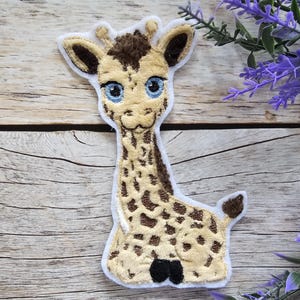 Könnte beinhalten: Ein gestickter Giraffenaufnäher mit cremefarbenem Körper und braunen Flecken, großen blauen Augen und dunkelbraunen Haarbüscheln auf dem Kopf. Der Aufnäher hat einen weißen Rand und zeigt die Giraffe in sitzender Position mit langem Hals.