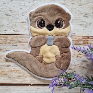 Könnte beinhalten: Ein gestickter Plüschaufnäher in Form eines braunen Otters mit großen dunklen Augen, einer cremefarbenen Brust und einem kleinen grauen Stein zwischen den Pfoten. Der Aufnäher hat einen weißen Filzrand und eine weiche, strukturierte Oberfläche.