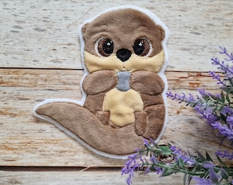 May include: A plush embroidered patch featuring a brown otter with large dark eyes, a cream-colored chest, and a small grey stone held between its paws. The patch has a white felt border and a soft, textured surface with detailed stitching.