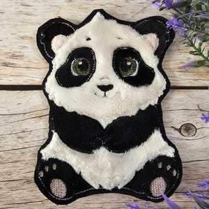 Könnte beinhalten: Ein Plüsch-Panda-Applikationen mit schwarz-weißem Fell. Der Panda hat eine schwarze Nase und schwarze Augen mit grünen Pupillen. Der Panda sitzt mit seinen Pfoten vor sich.