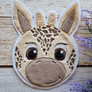 Könnte beinhalten: Applikation eines Giraffengesichts aus Plüsch mit weißem Rand. Die Giraffe hat braune und beige Flecken, große braune Augen und eine weiche, beige Schnauze. Die Ohren sind beige mit dunklerem braunen Innenfutter. Für Kinderbastelprojekte.
