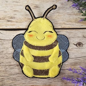 Può includere: Un'ape ricamata di colore giallo e marrone con un viso sorridente e ali blu. L'ape è seduta su una superficie di legno.