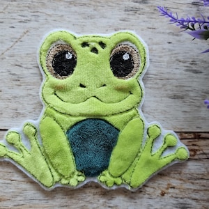 Könnte beinhalten: Ein grüner Plüschfrosch mit großen, schwarzen Augen und einem weißen Umriss. Der Frosch sitzt mit gekreuzten Beinen und hat einen grün-blauen Bauch.
