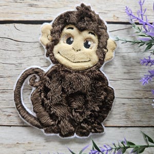 May include: A dark brown embroidered monkey patch with a light beige face, ears, and hands. The monkey has a textured fur-like stitch pattern, a curled tail, and a white border outline. The design features black embroidered eyes and a small smile.