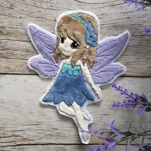 Può includere: Applique di fata in feltro viola e blu con contorno bianco. La fata ha i capelli castani con una fascia blu e un vestito blu chiaro.