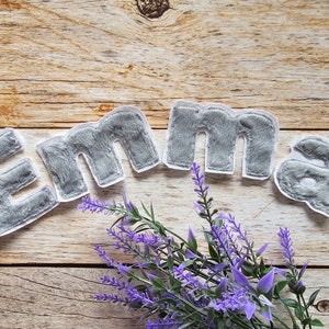 Könnte beinhalten: Graue Filzbuchstaben, die den Namen "Emma" auf einem Holzgrund mit lila Blumen buchstabieren.