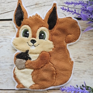Può includere: Toppa a forma di scoiattolo di peluche con pelliccia marrone, orecchie nere e coda soffice. Lo scoiattolo tiene in mano una ghianda nera e marrone. La toppa ha un bordo bianco e occhi verdi.