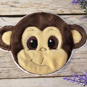 Op de afbeelding: Een bruin en geel pluche apen gezicht met grote ogen en een glimlach. De aap is geborduurd met witte stiksels.